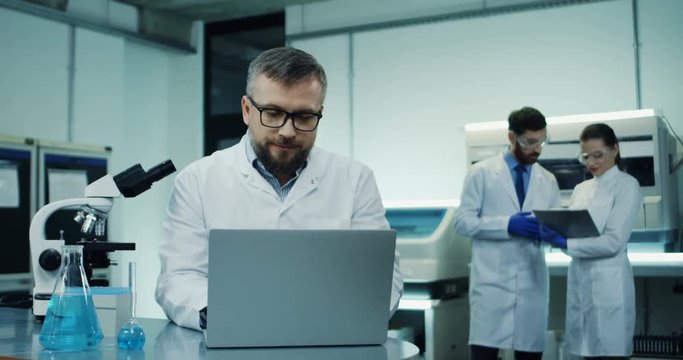 Close up of the Caucasian man in glasses, medical researcher, looking in the microscope while doing some analysis and typing them in the laptop computer at the laboratory.