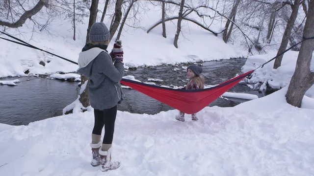 Girl swinging in hammock posing for picture in the snow as her sister takes photos of her next to river.