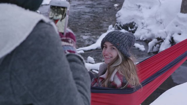 Girl smiling for photo as she sits in hammock outside in the snow during winter as her sister takes man pictures.