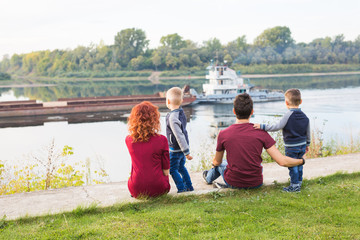 Parenthood, childhood and nature concept - Family sitting on the green ground and looking at small boat