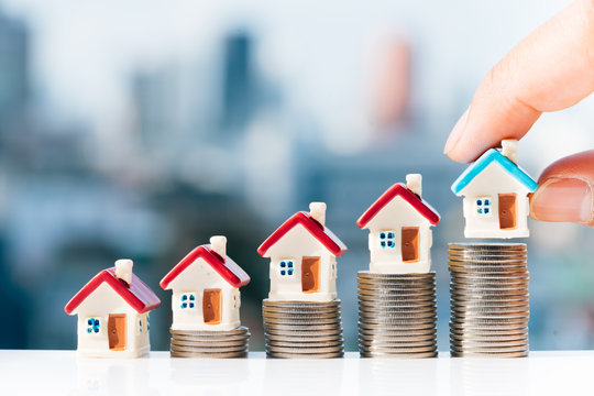 Concept for property ladder, mortgage and real estate investment. Man's hand putting blue house model on top of coins stack with city backgrounds.