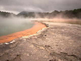 Volcanic rock pool with layers of gray, green, and orange rock with steam rising from water and trees and hills in background under grey, cloudy sky