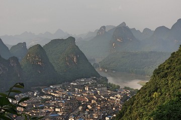 Limestone karst peaks in Yangshuo, Guangxi, China with town low-rise and high-rise buildings in valley and river underneath blue sky with hazy smog in air