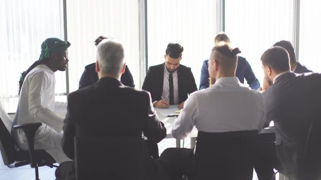 Arabian investor in white national candura and gutra on head discussing company financial strategy with his multirucial male managers during meeting at desk in office.