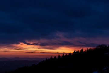 Trees silhouettes against a beautifully colored sky at dusk, with mountains layers in the background