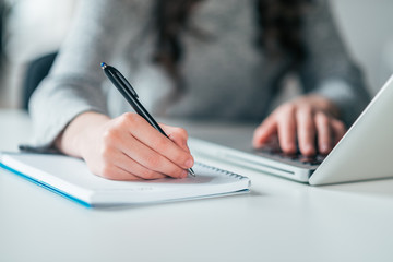 Front view of female student writing in notebook and using laptop, close-up.