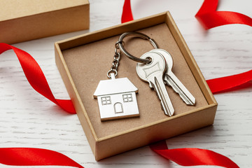 Keychain house and keys with red ribbon and gift box on white wooden background.