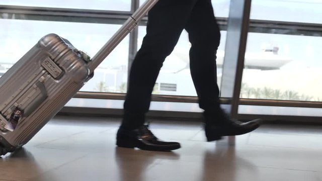 Silver trolley suitcase rolled across airport by businessman. Legs tracking shot