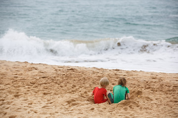 Brother and sister playing with sand and water on a tropical beach, dressed in protective wetsuit.