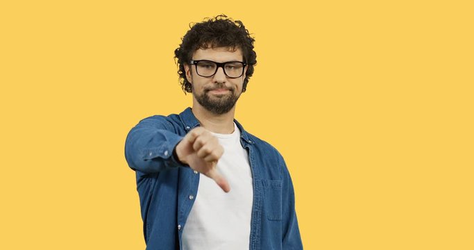 Caucasian young man in glasses with curly hair giving his thumb down while standing on the yellow screen background.