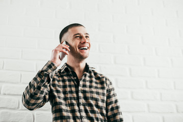 A young man stands with a mobile phone and talks smiles and laughs in a plaid shirt