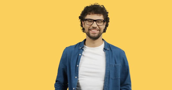 Portrait shot of the attractive smiled man with curly hair and in glasses posing cheerfully to the camera on the yellow background because of the winning or good result.