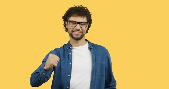 Young attractive Caucasian man in glasses standing in front of the camera and doing yes, well done gesture with a hand. Yellow back screen.