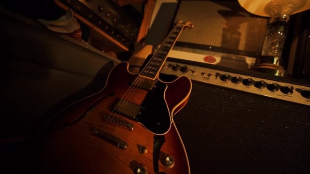 A hand held camera tilts up from a garage band practice space basking in warm light, with amplifiers, guitars, microphones, and an old lamp.