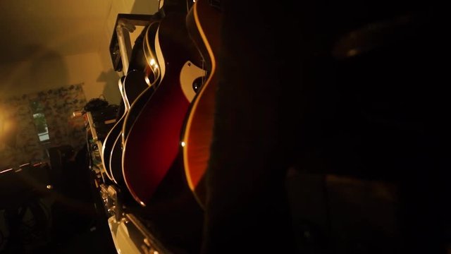 A hand held camera tilts down a row of guitars in a garage band practice space basking in warm light.
