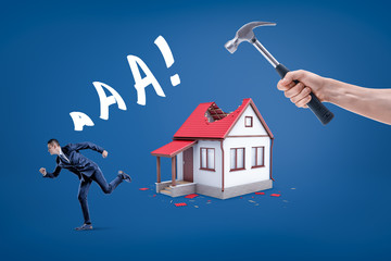 A hand holding a hammer breaking small house's roof with a businessman running away screaming.