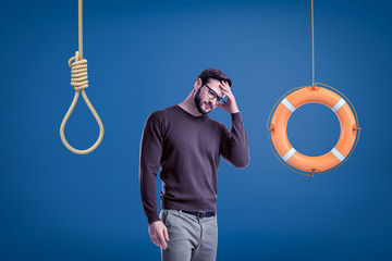 Man in casual outfit standing half-turn with hand on forehead as if choosing between lifebuoy hanging on rope and hangman's noose.