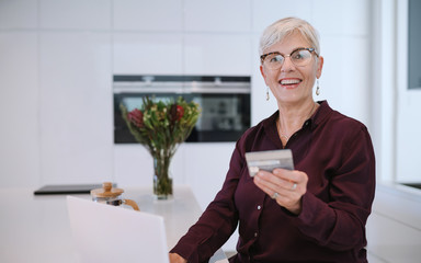 Happy senior woman making online payments of bill using laptop. Smiling mature woman shopping online with credit card. Pensioner holding credit card for internet banking and looking into the camera