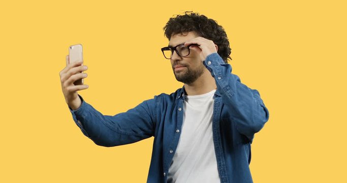 Handsome Caucasian young man with curly hair in glasses making selfie on the smartphone while standing on the yellow screen back.