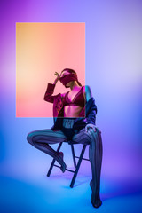 Beautiful woman with long hair in black underwear, pantyhose, jacket and scarf, covering the mouth posing in attractive position in colorful bright neon blue and pink lights in studio. Fashion concept