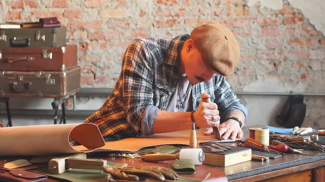 Handsome young shoemaker, wearing a shirt and a cap, cutting leather in workshop with special knife