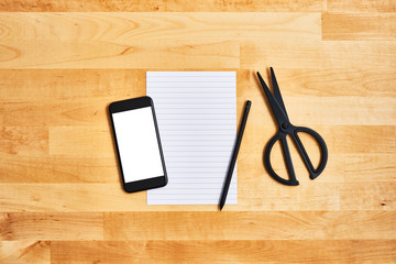 An empty sheet of paper with black smartphone, pencil and scissors on yellow wooden table. School concept. Top view. Copy space for text.