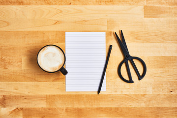 Cup of coffee or cappuccino with an empty sheet of paper, scissors and pencil on yellow wooden table. Top view. Copy space for text.