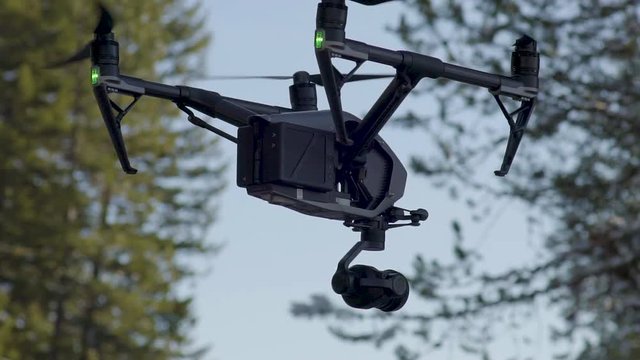 A slow motion close up of a DJI Inspire 2 quadcopter drone hovering in the air with blue skies and green trees in the background.