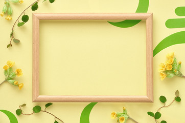 Wooden frame decorated with yellow primrose flowers and leaves, flat lay on yellow paper, copy-space