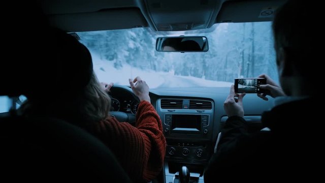 Young couple of travelers and adventurers drives small car or camping van through winding snow covered mountain road on cold winter vacation day. Man makes photos with smartphone of views and girl