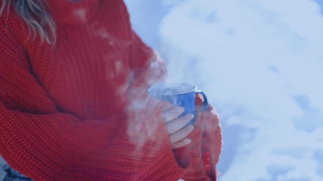 Unrecognizable woman in red knit woolen sweater holds a cup with steaming hot coffee or tea. Apres ski winter lifestyle or camping trip in harsh conditions in negative temperatures