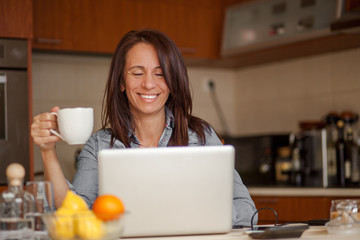 Obraz na płótnie Canvas Smiling mid adult woman drinking coffee and using laptop in kitchen