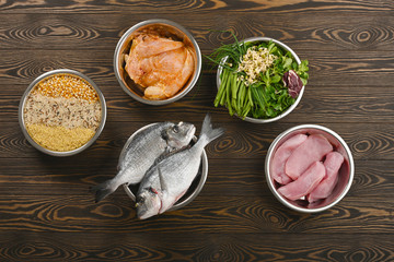 Panorama banner with healthy pet food ingredients with chopped raw turkey, fish, groats, greens and grains in individual bowls on brown wooden background. Flat lay.