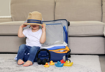 Cute funny little baby boy siiting in blue suitcase with hat on his eyes, packed for vacation full of clothes ready for traveling. Vacation with child.