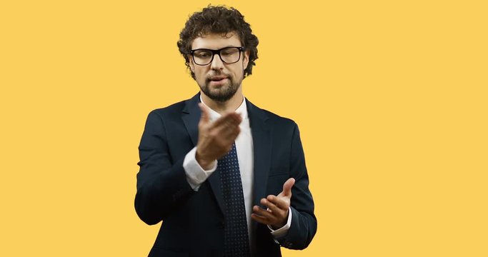 Good looking Caucasian businessman in glasses, suit and tie standing on the yellow background and explaining something with gestures.