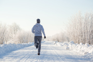 Young, adult alone man in tracksuit and hat running on snowy road outside of city. Enjoying sport in sunny winter morning. Daily active lifestyle. Life motion. Back view. 