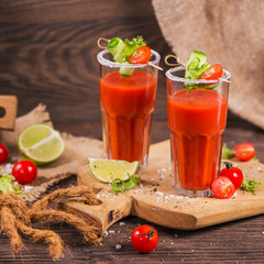 Two glasses of fresh organic tomato juice decorated with raw tomatoes, cucumber and leaves on a rustic wooden cutting board