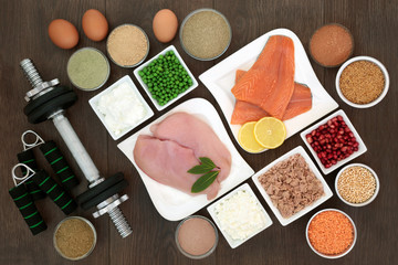 Body building dumbbell weights and hand grippers with high protein lean meat, fish and dairy, dietary supplement powders, vegetables, fruit and grains. Top view on oak wood table.