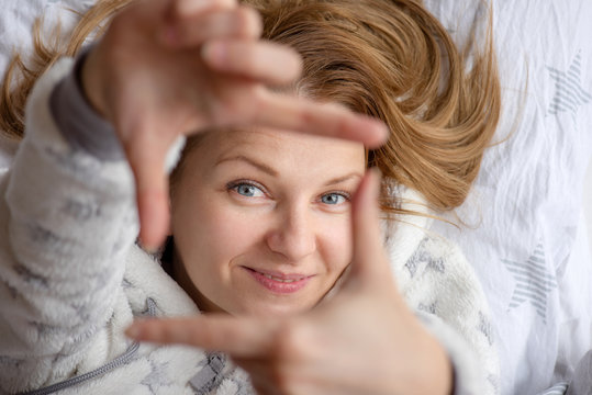 Top view Portrait of Pretty Girl lying in Bed in Pajamas. Young Woman having a good sleep with open eyes, smiling making frame with hands and fingers with happy face.