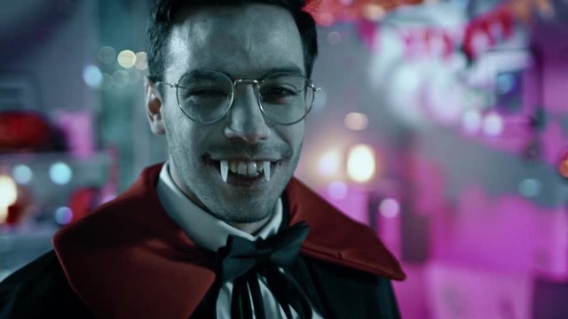 Halloween Costume Party: Count Dracula Revealing Himself From Under The Cape and Shows His Deadly Fangs. In the Background Thematically Decorated Room with Scary Stuff. Bloody Red Colors