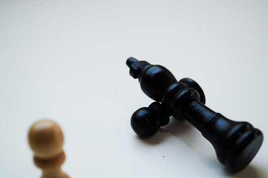 Black King and his pawn thrown defeated in front of white pawn. Photograph in white background iluminated from behind.