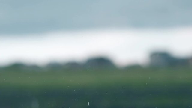 view though car windshield rain falls on green blurry field against building silhouettes under sky with big gray cloud