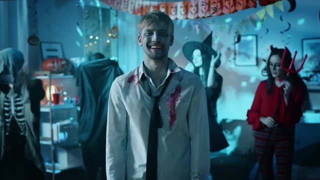 Halloween Costume Party: Creepy Zombie Wearing Torn Bloody Suit Laughs. In the Background Beautiful Witch, Gorgeous She Devil and Scary Death and Count Dracula Dancing in the Decorated Room