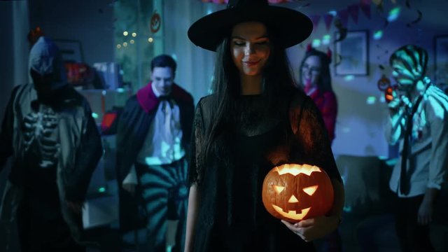 Halloween Costume Party: Gorgeous Witch Wearing Dress Holds Burning Pumpkin and Dances Seductively. Background: Beautiful She Devil, Scary Death, Count Dracula, Zombie Dancing in the Decorated Room