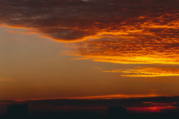 Cityscape with vivid fiery dawn. Amazing warm dramatic cloudy sky above dark silhouettes of city building roofs. Orange sunlight. Atmospheric background of sunrise in overcast weather. Copy space.