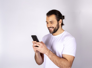 Handsome man in a white T-shirt on a white background, smiling, rejoices, in the hands of a smartphone, writes SMS