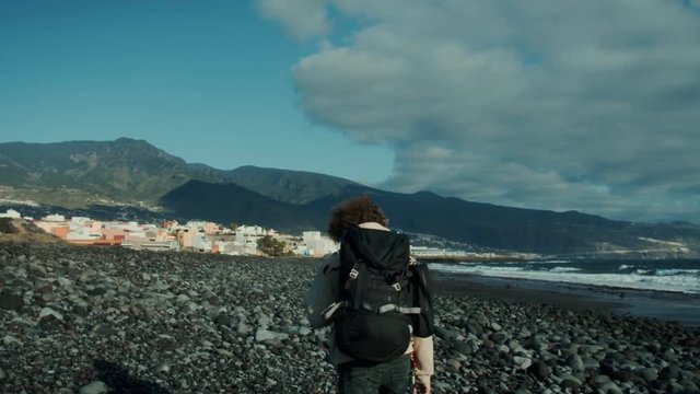 Cool and serious middle aged man with beard and curly hair, wears backpack and sunglasses, walks on empty beach, does a long dramatic stare at camera, trips and falls in funny fail way