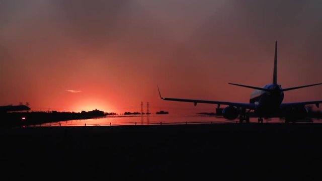 An airplane waits on a runway as another airplane lands in the background against a the sunset