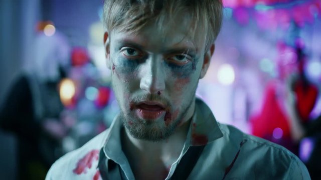 Halloween Costume Party: Portrait of Brain Dead Zombie Wearing Bloody Suit Smiles Creepily. In the Background Monsters Having Fun and Dancing in the Decorated Room