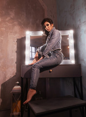 gorgeous sexy woman with bright makeup in dark blue denim wear sitting on table near professional lighting mirror looking at camera with passion.